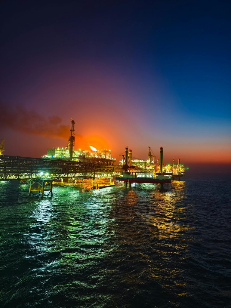 Illuminated offshore oil rig in the ocean during a vibrant sunset, reflecting on the water.