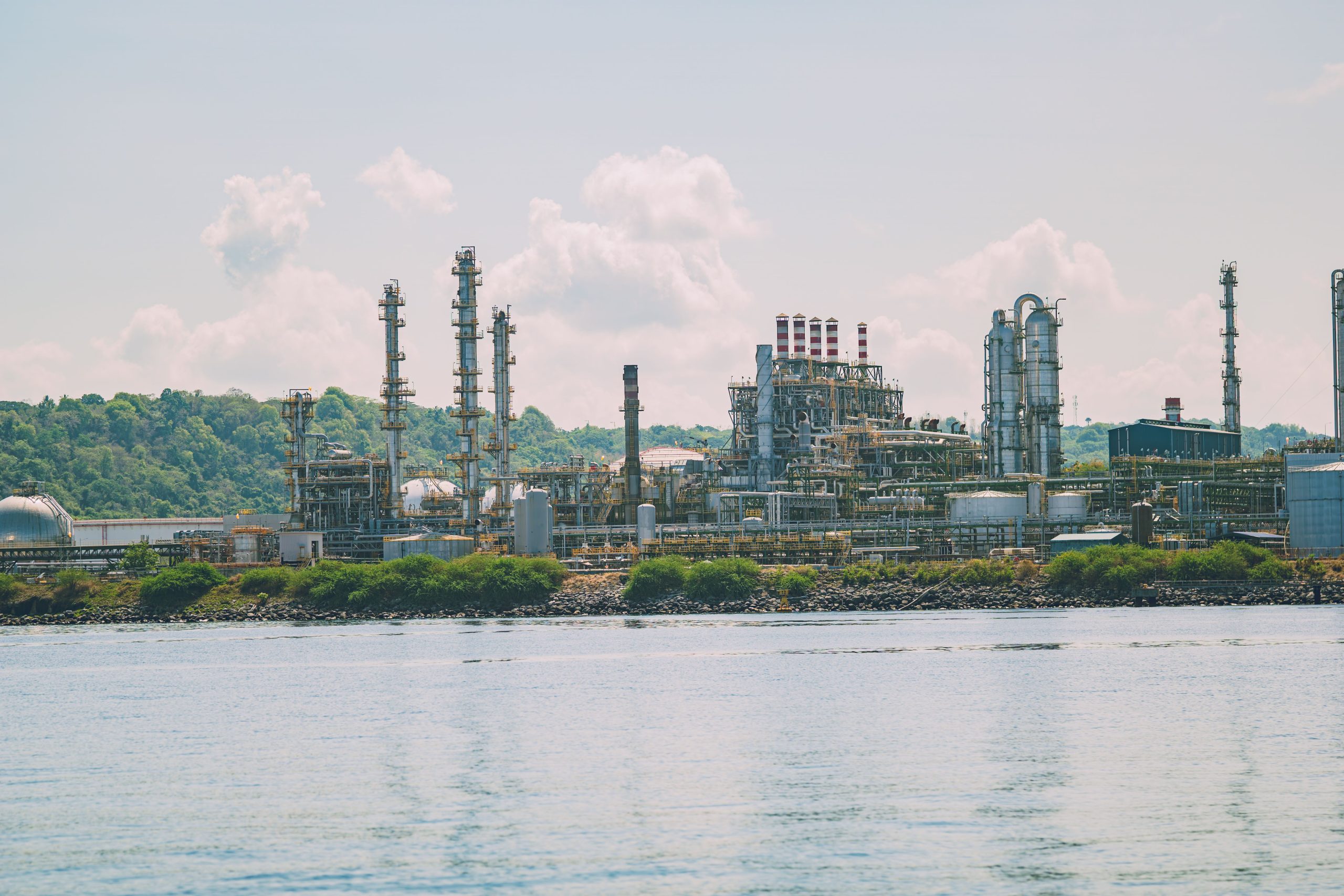 A large oil refinery situated by the water with bright green foliage in the background; clear blue skies and clouds overhead.